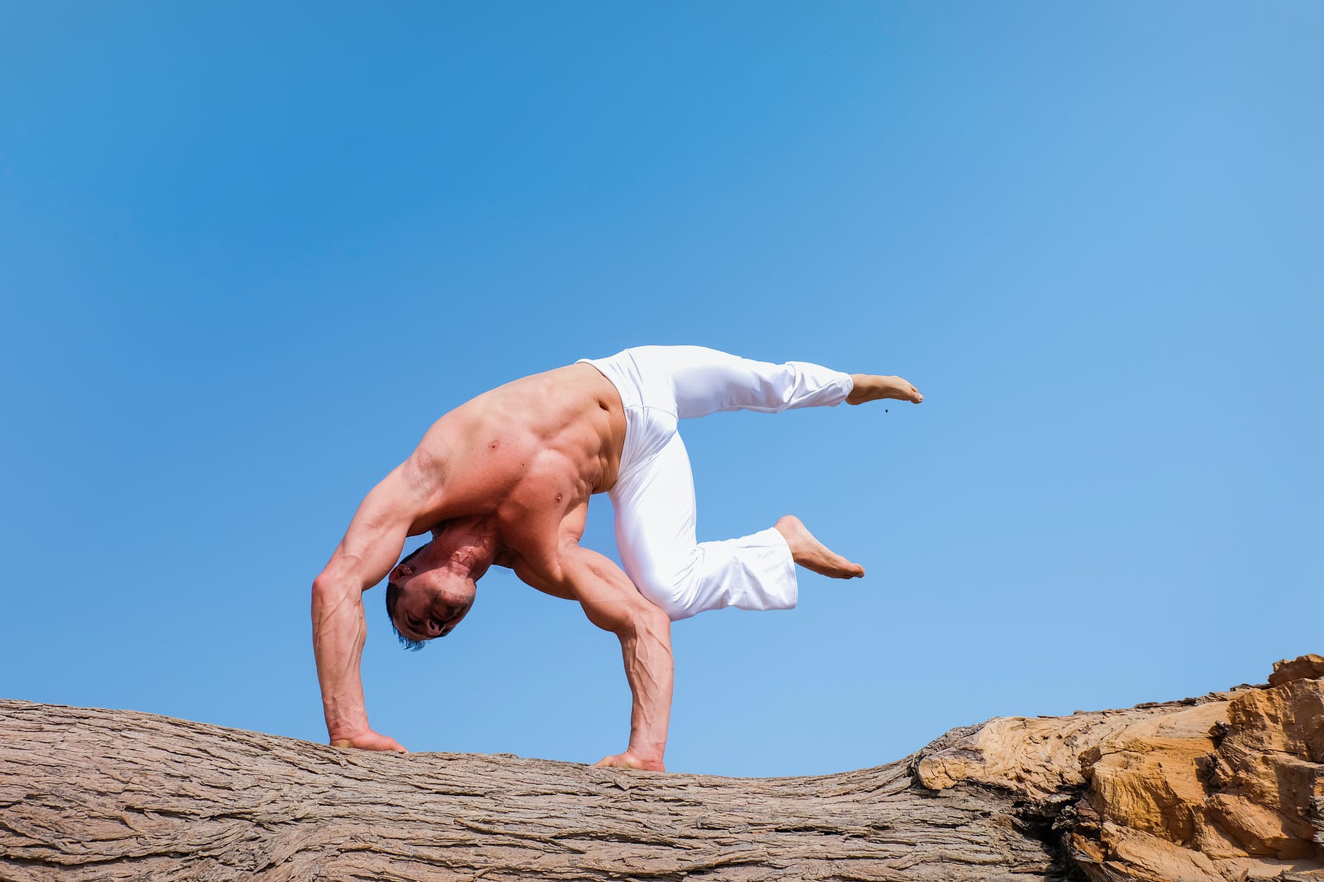 man wearing white pants under blue sky, doing an inverted yoga pose