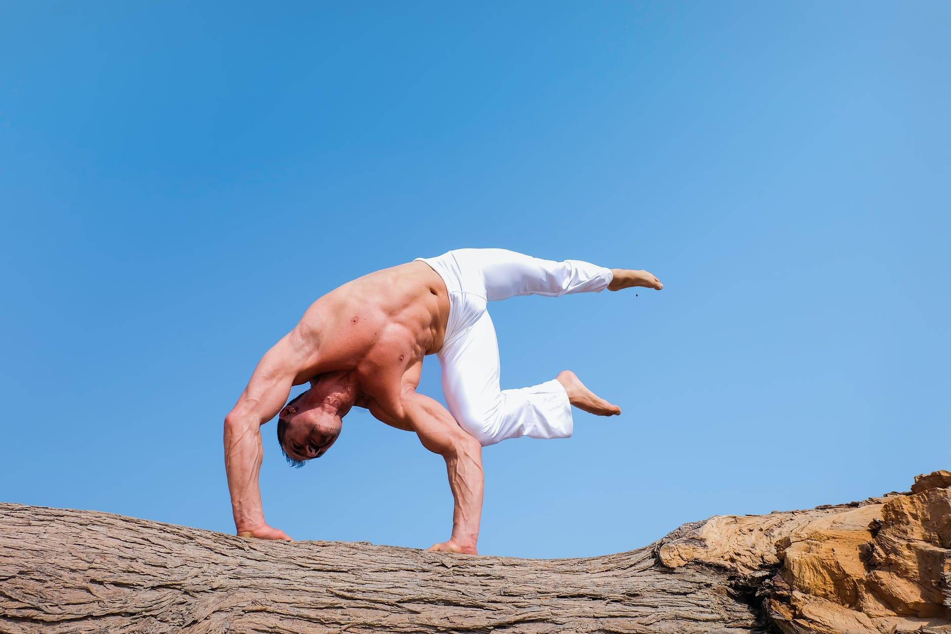 man wearing white pants under blue sky, doing an inverted yoga pose